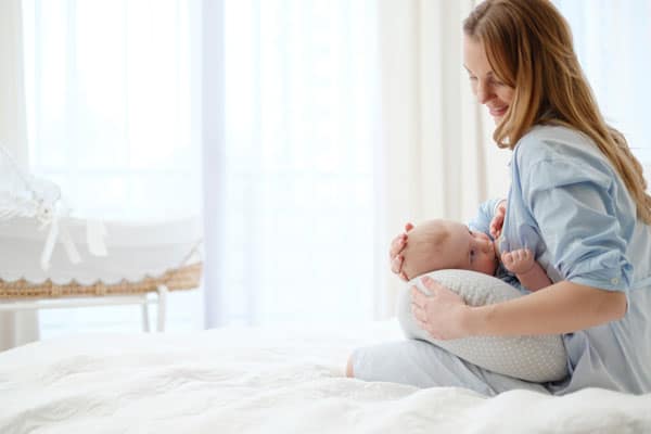 mom nursing newborn baby with breastfeeding pillow on bed with moses basket in the background